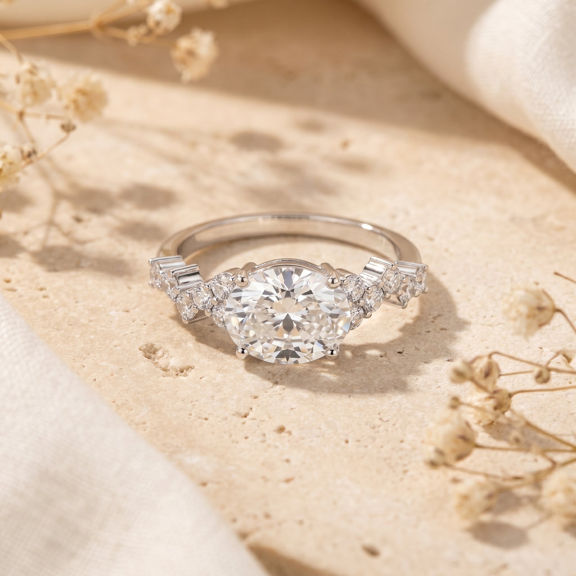 Silver oval moissanite promise ring with cluster pavé side stones on a stone surface with dried flowers.