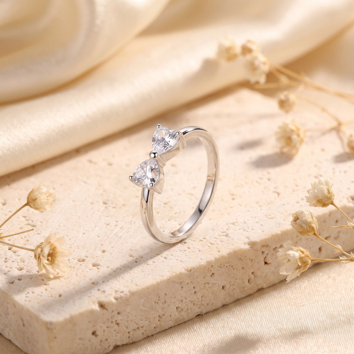 Angled view of a silver ring with two heart-cut clear zircons forming a bow, resting on a stone block.
