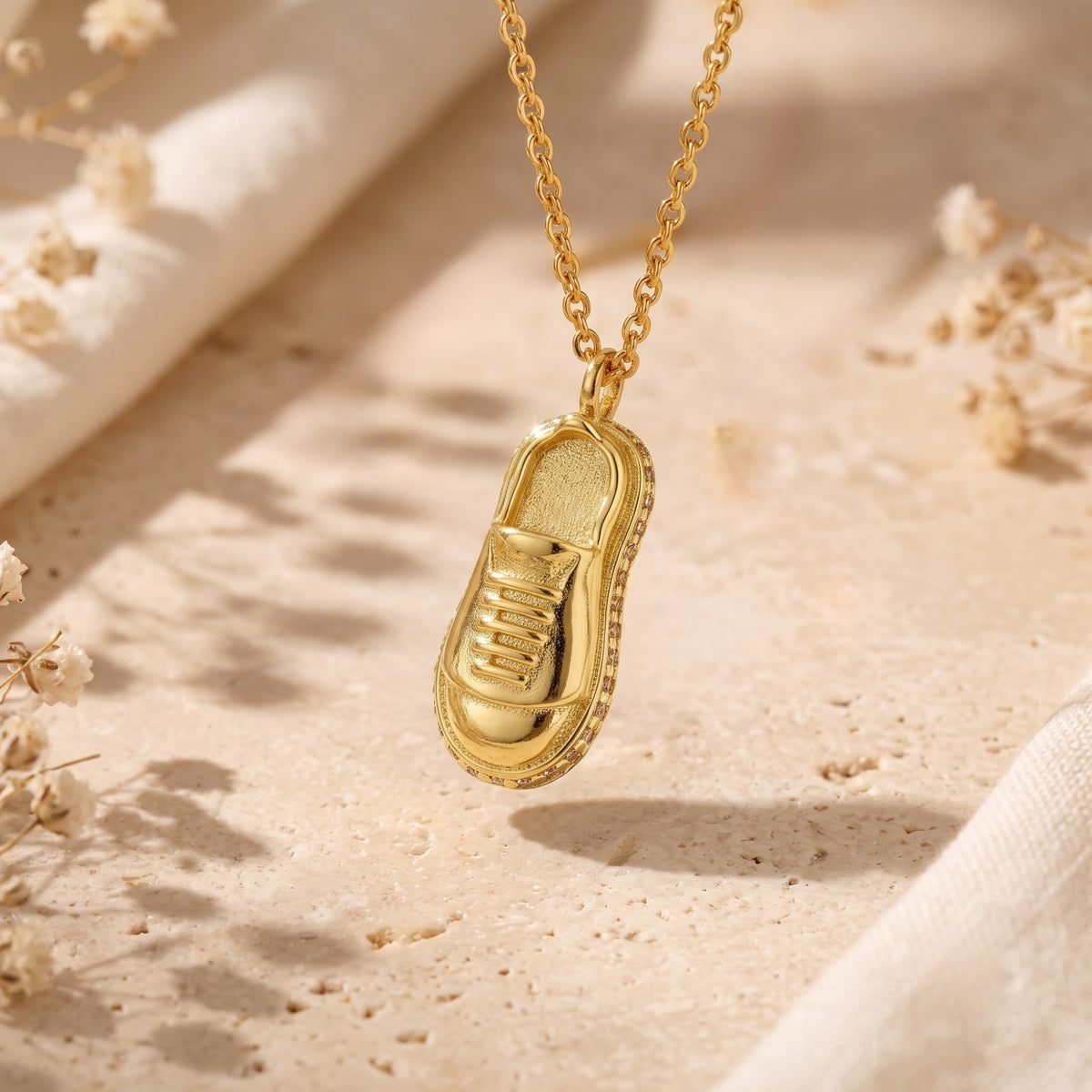 Gold sneaker pendant with stone-embellished sole hanging vertically against a warm stone backdrop.