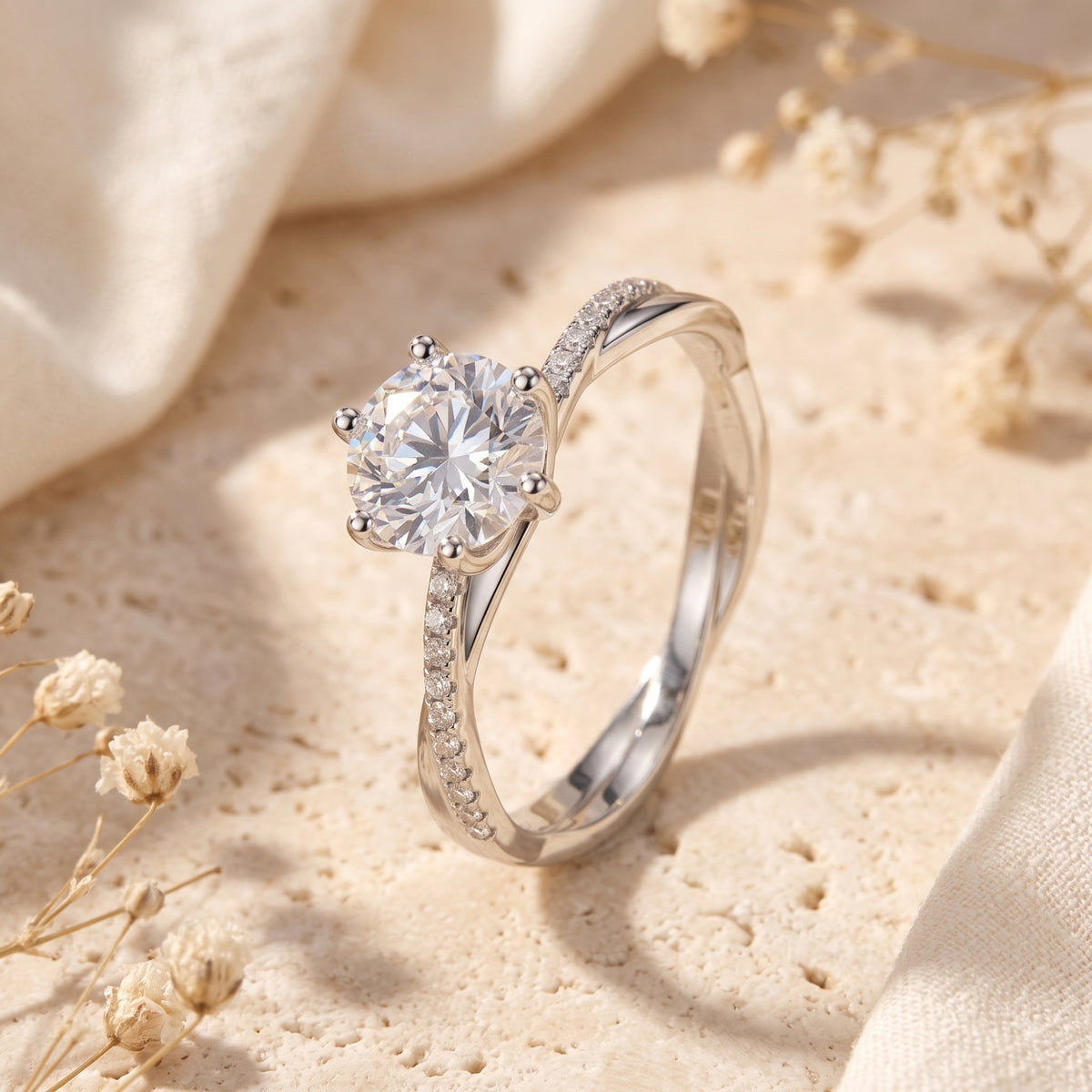 Angled silver crossover ring with a round clear center stone on a beige stone surface with dried flowers.