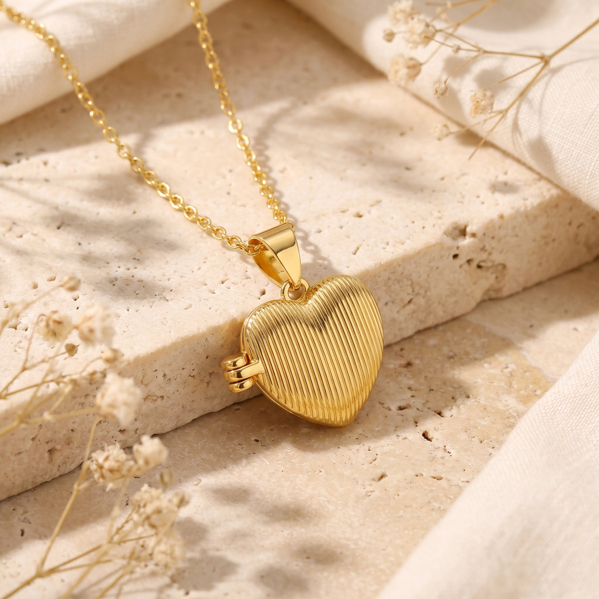 Gold-tone ribbed heart pendant locket on a chain, resting on a textured stone surface with dried flowers.