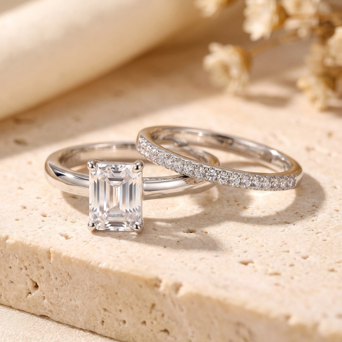 Two silver rings on a stone surface: one with an emerald-cut center stone and one with a pavé band.