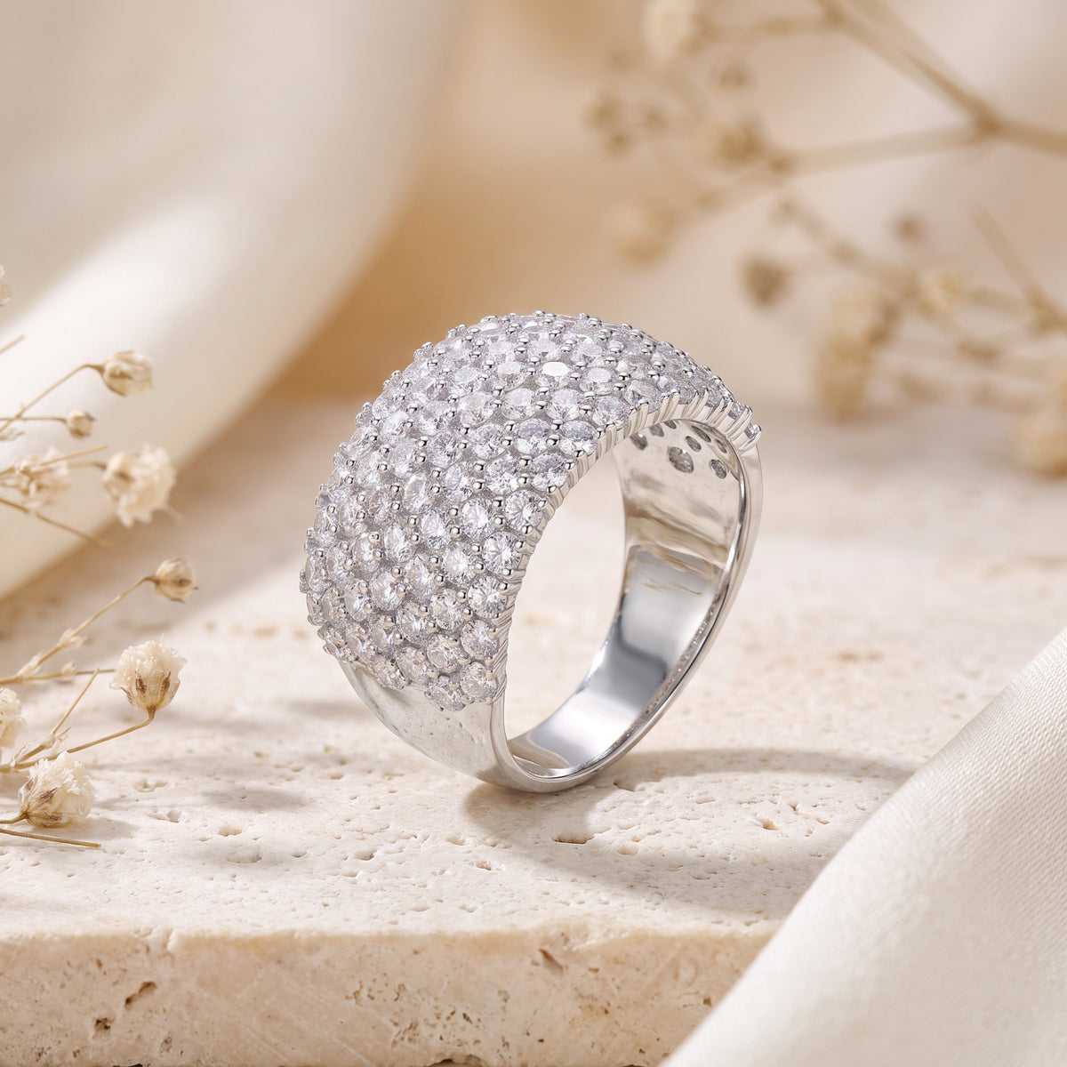 Wide silver dome band with multi-row clear pave stones, standing at an angle on a textured beige stone surface.