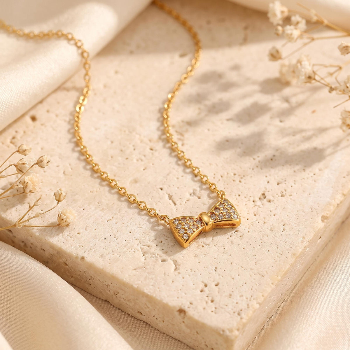 Gold-tone bow pendant necklace set with pave cubic zirconia, resting on a textured stone block with silk fabric.