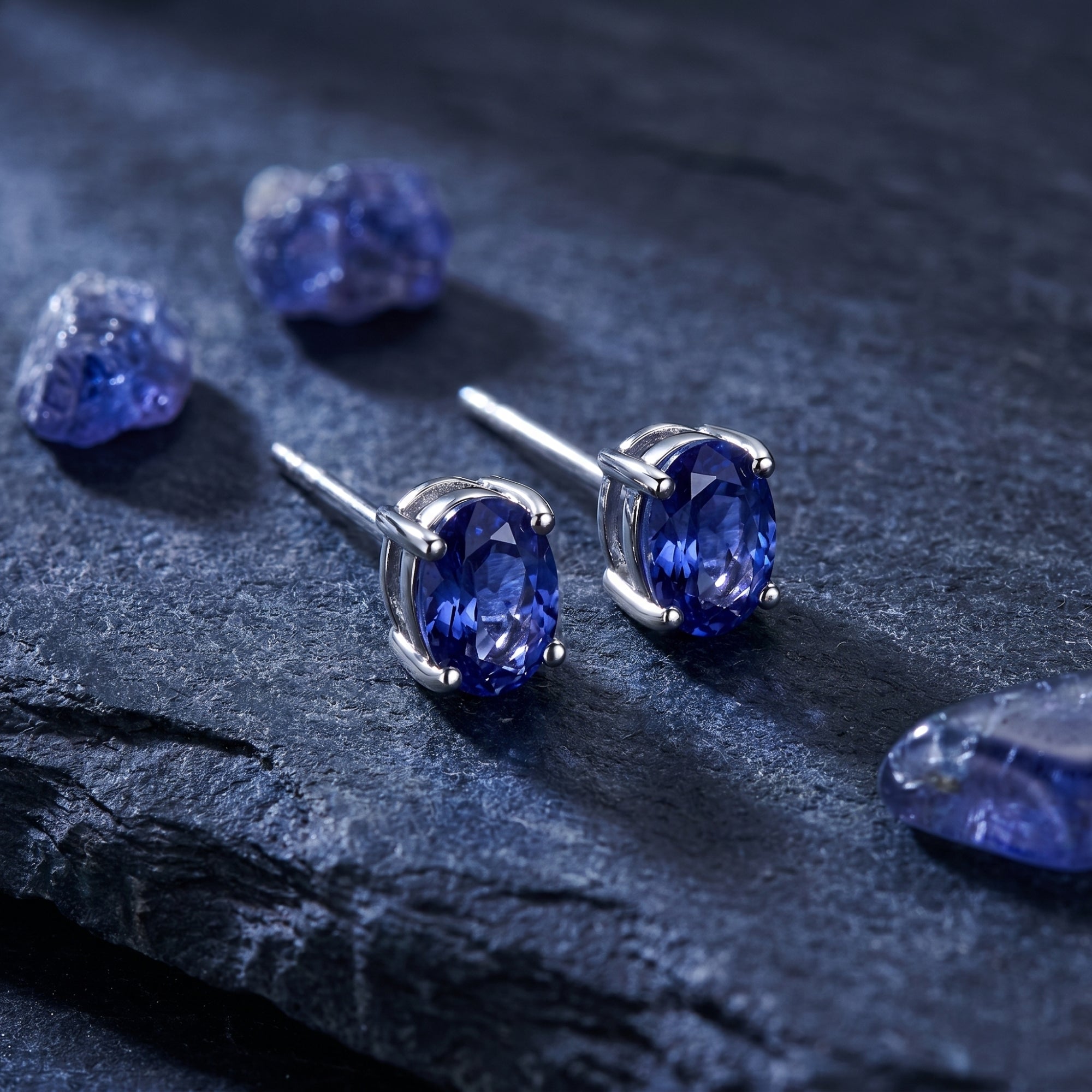 Oval cut natural tanzanite stud earrings in 18K white gold on a dark stone surface with raw gemstones.