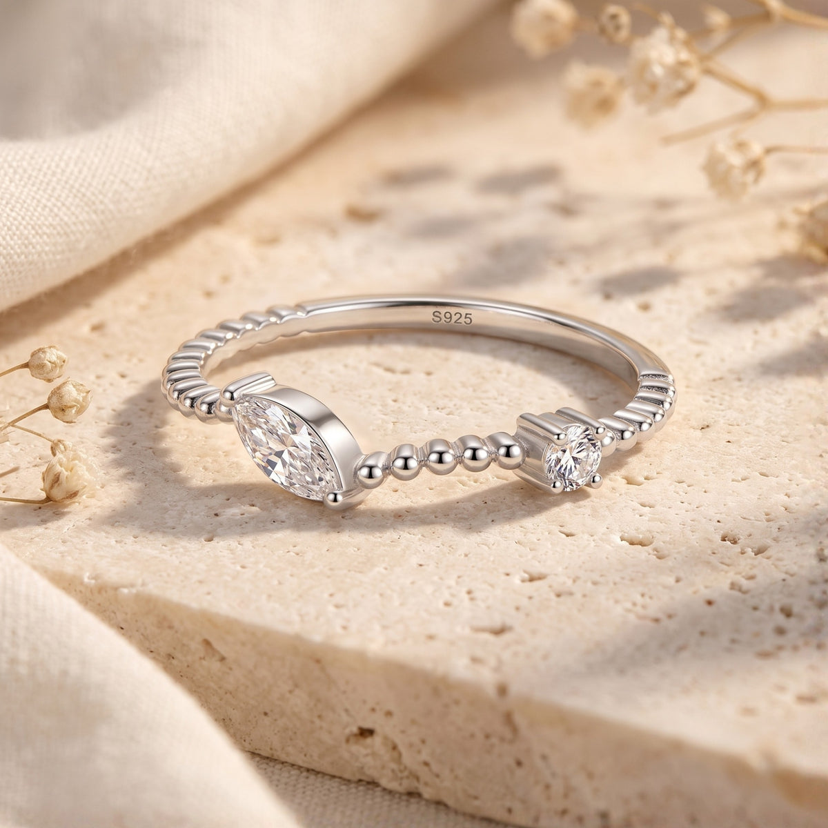 Silver beaded bypass ring with marquise and round clear stones resting on a textured beige stone slab.