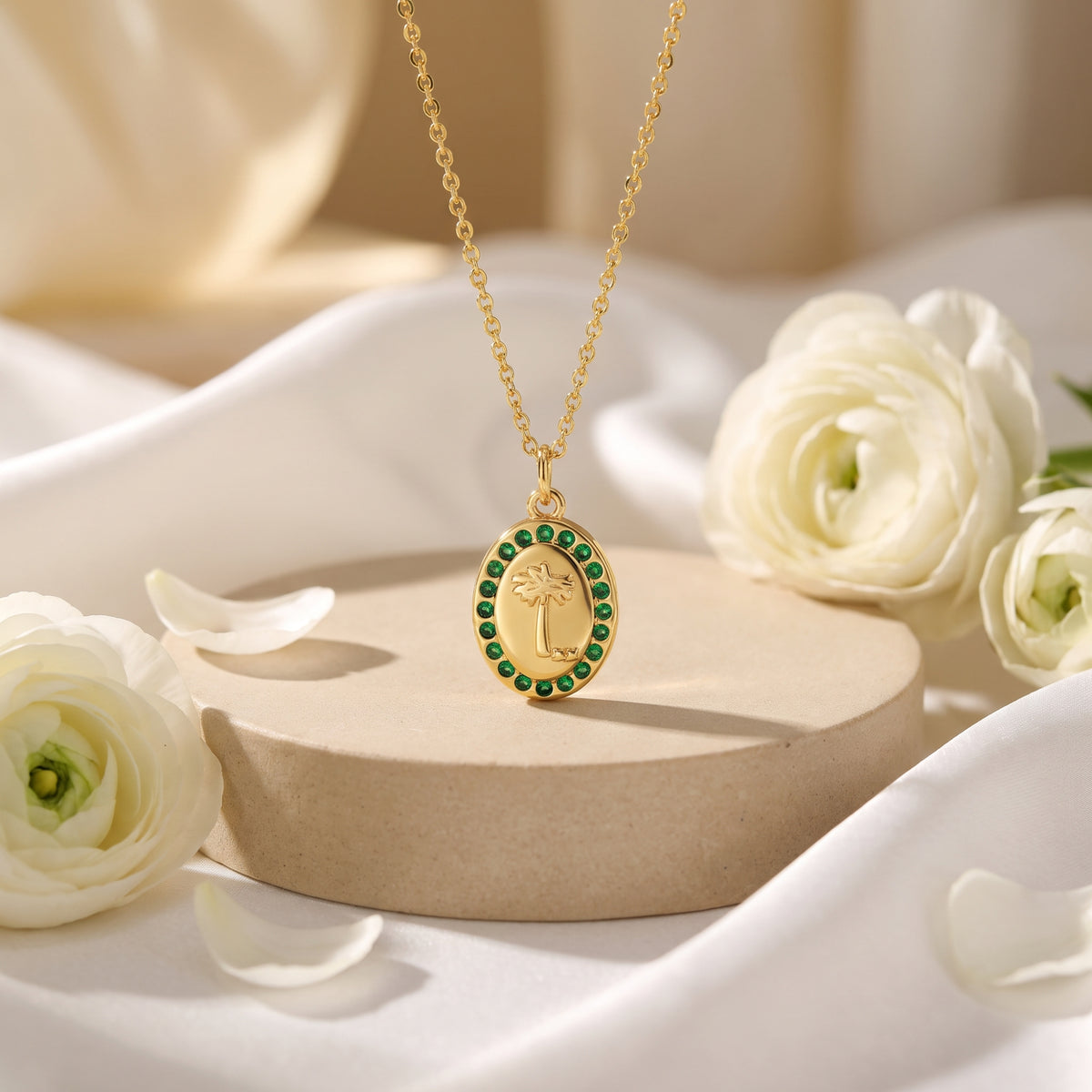 Gold-tone oval palm medallion necklace with green crystals on a beige pedestal surrounded by white flowers.