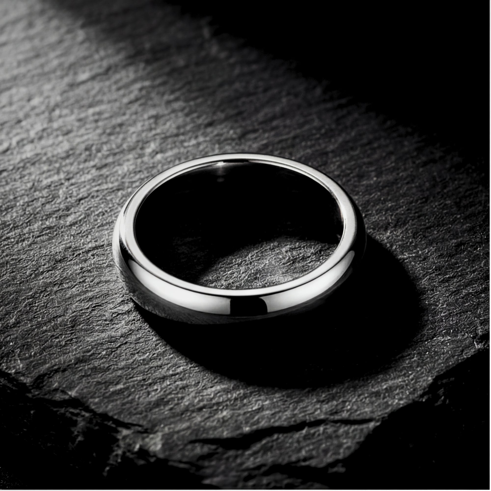 The Minimalist Ring, a silver 5mm stainless steel band, lying on a dark textured stone background.