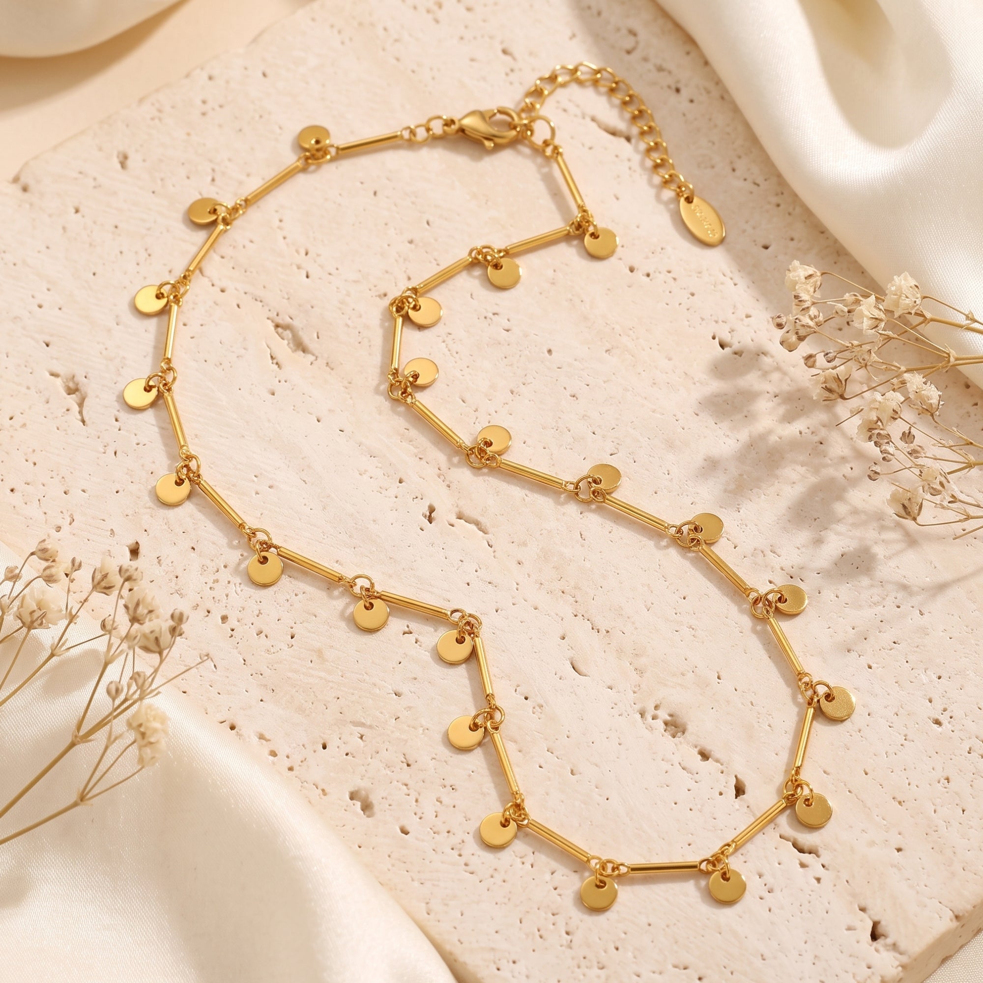 Gold disc drop necklace on a stone surface with floral accents.