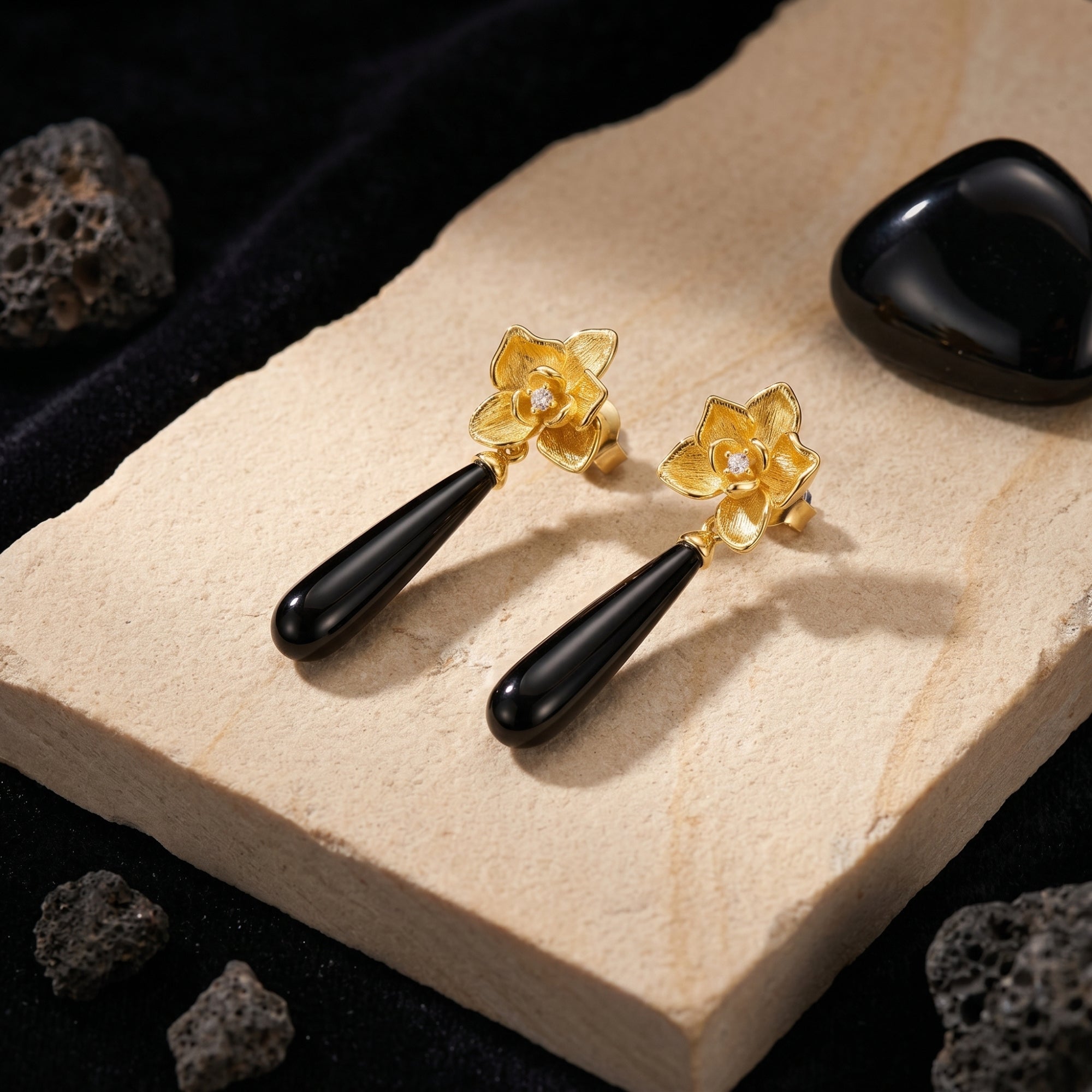 Angled view of 18K gold plated Calla floral drop earrings with elongated black onyx stones on a stone slab.