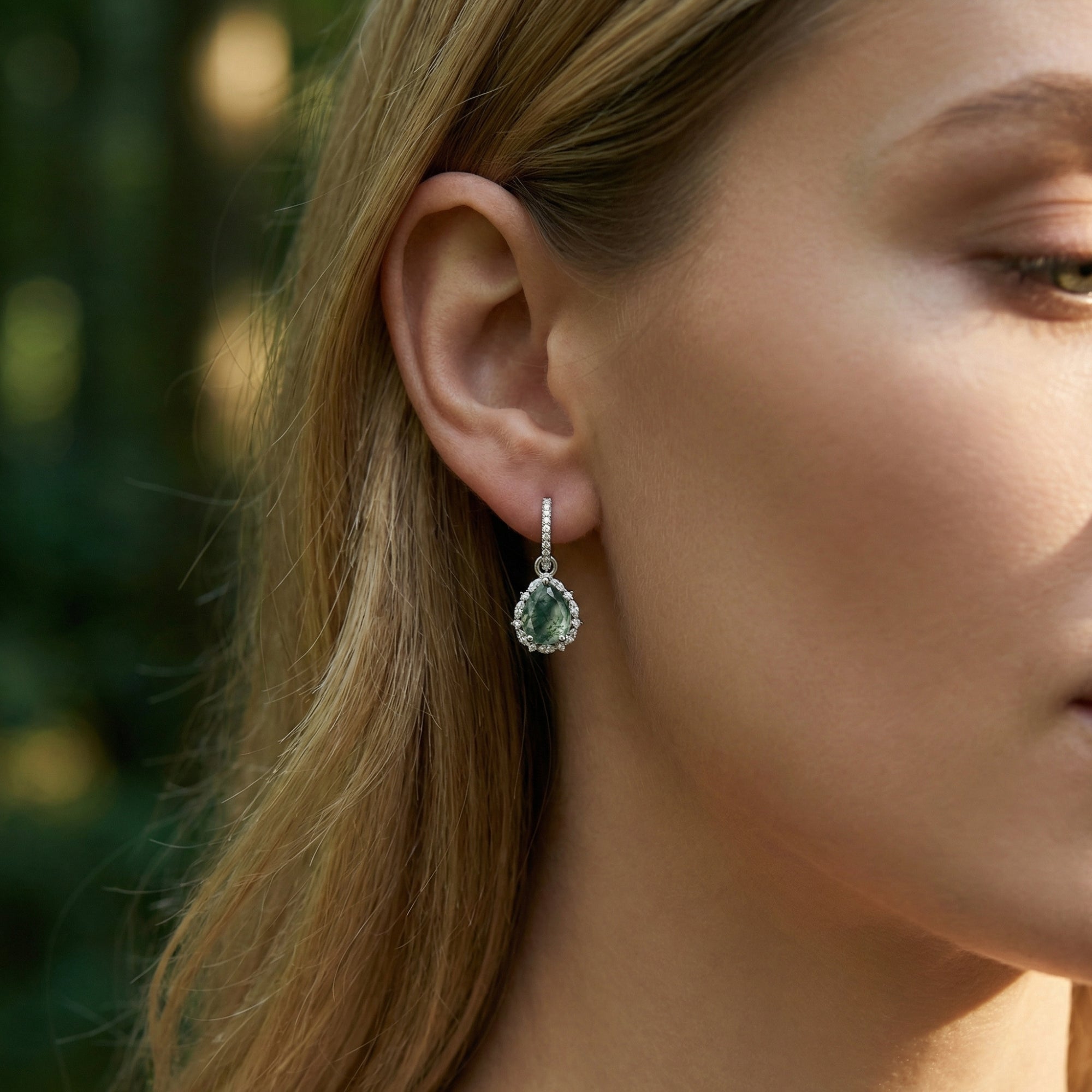 Side view of a model wearing a sterling silver teardrop moss agate halo earring against a soft forest background.