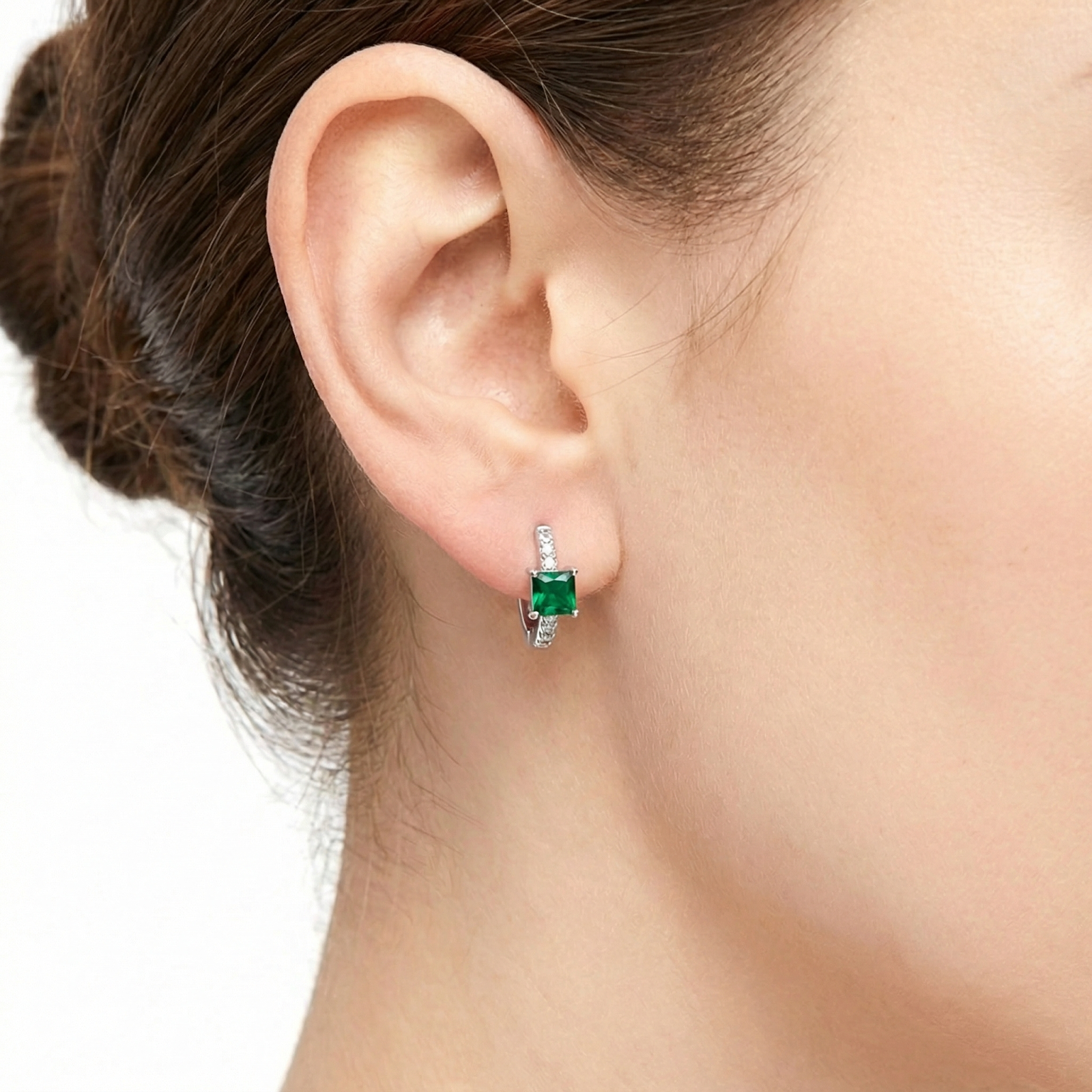 Close-up of a sterling silver huggie earring with a square-cut green simulated emerald worn on an ear.