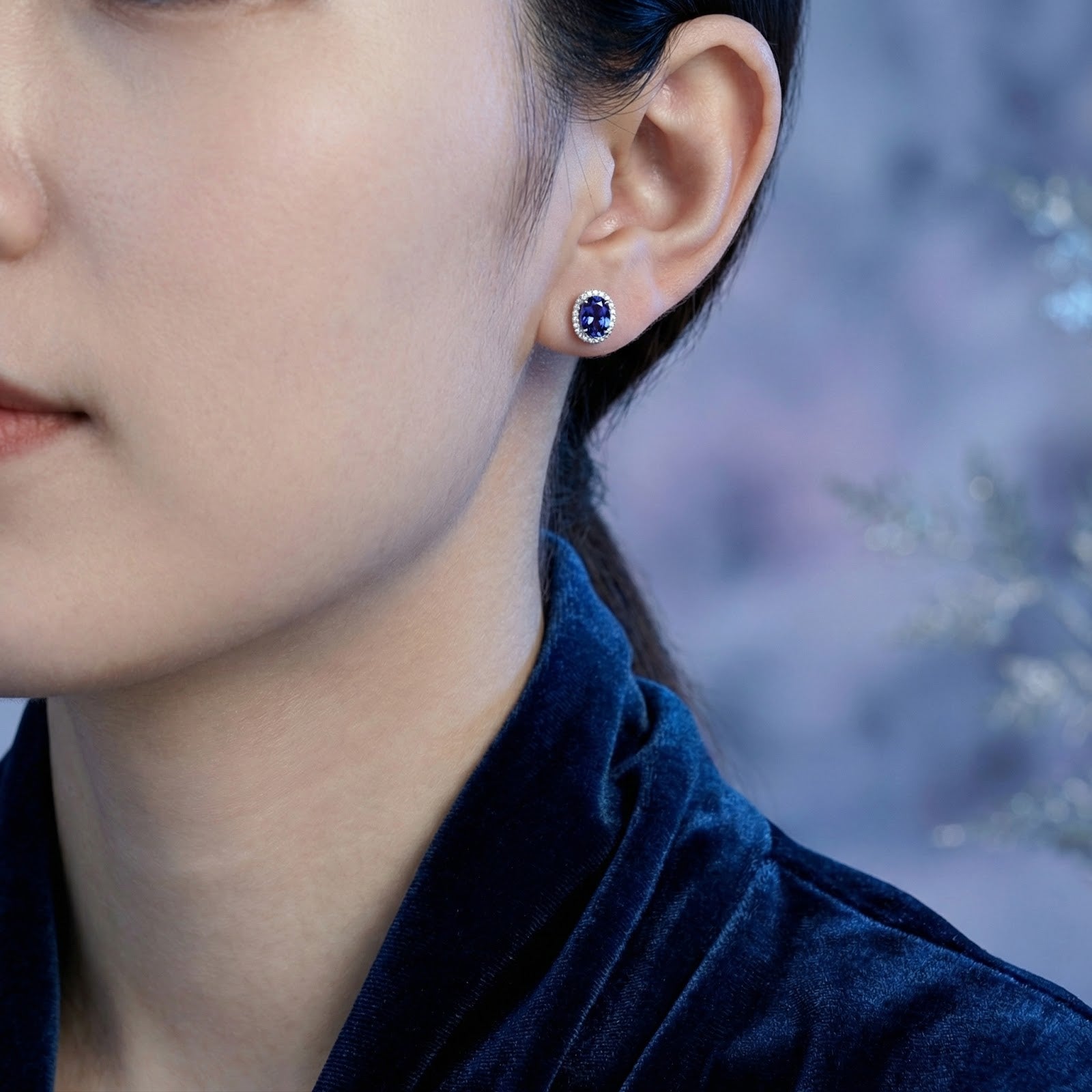 Woman wearing an oval simulated tanzanite halo stud earring in sterling silver.