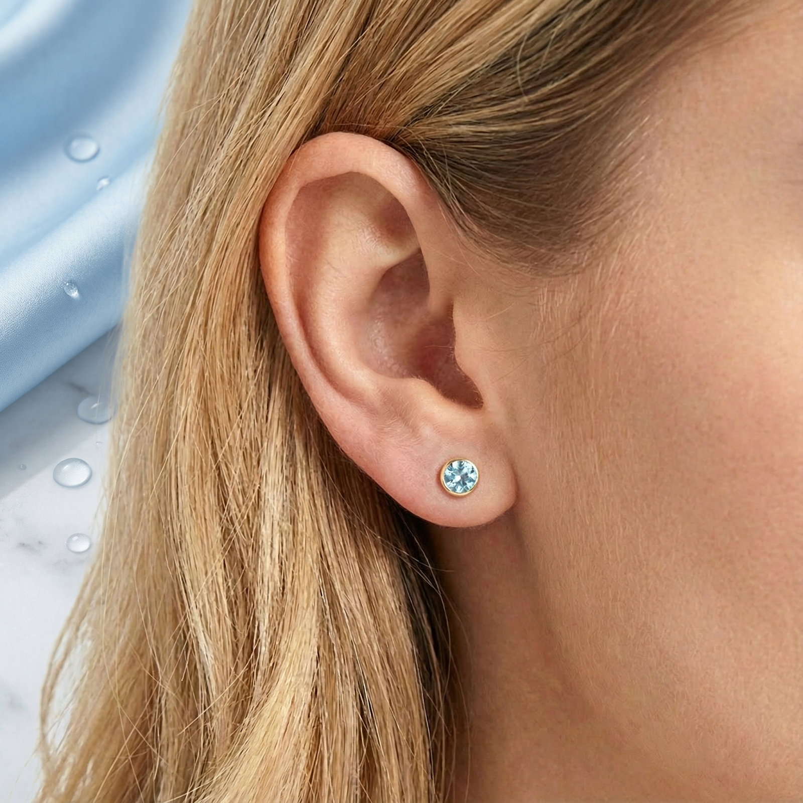 Close-up of a round simulated aquamarine stud earring in 18K gold vermeil worn on an ear.