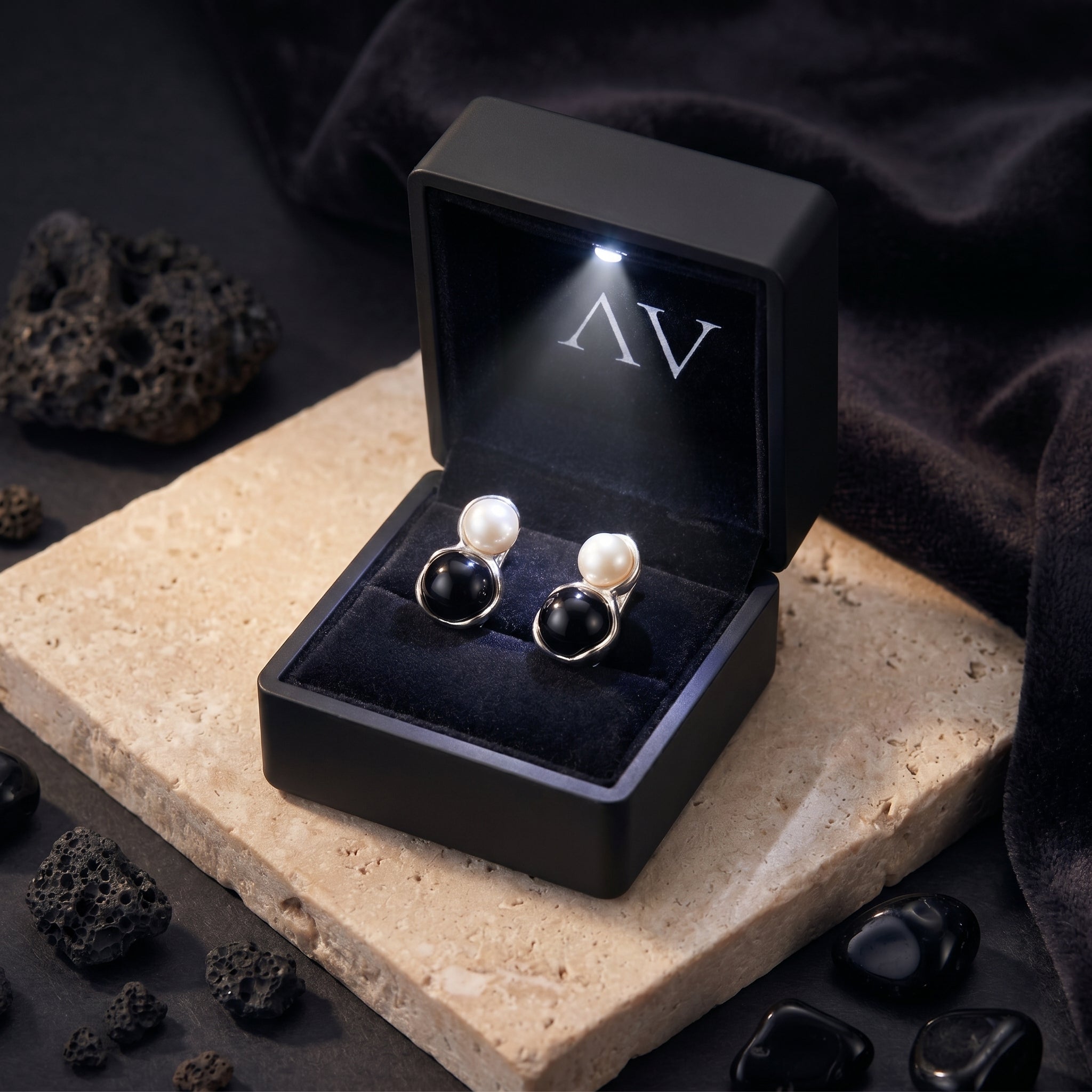 Silver pearl and black onyx drop earrings inside a lighted black jewelry gift box on a stone surface.