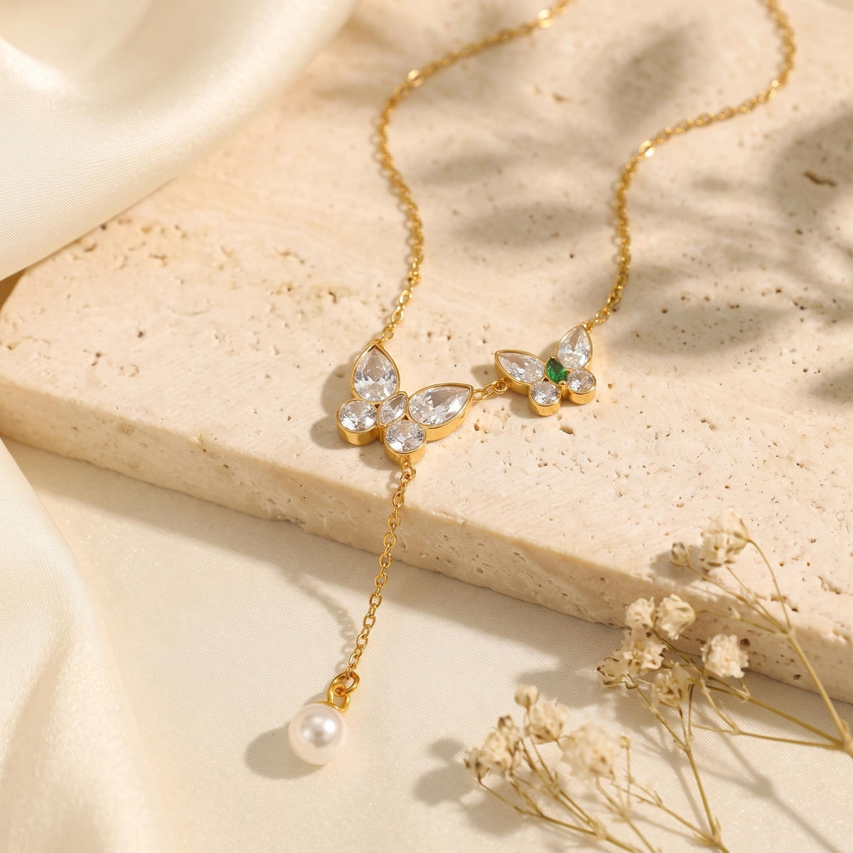 Gold-tone butterfly lariat necklace with clear stones, green accent, and dangling pearl on a textured beige surface.