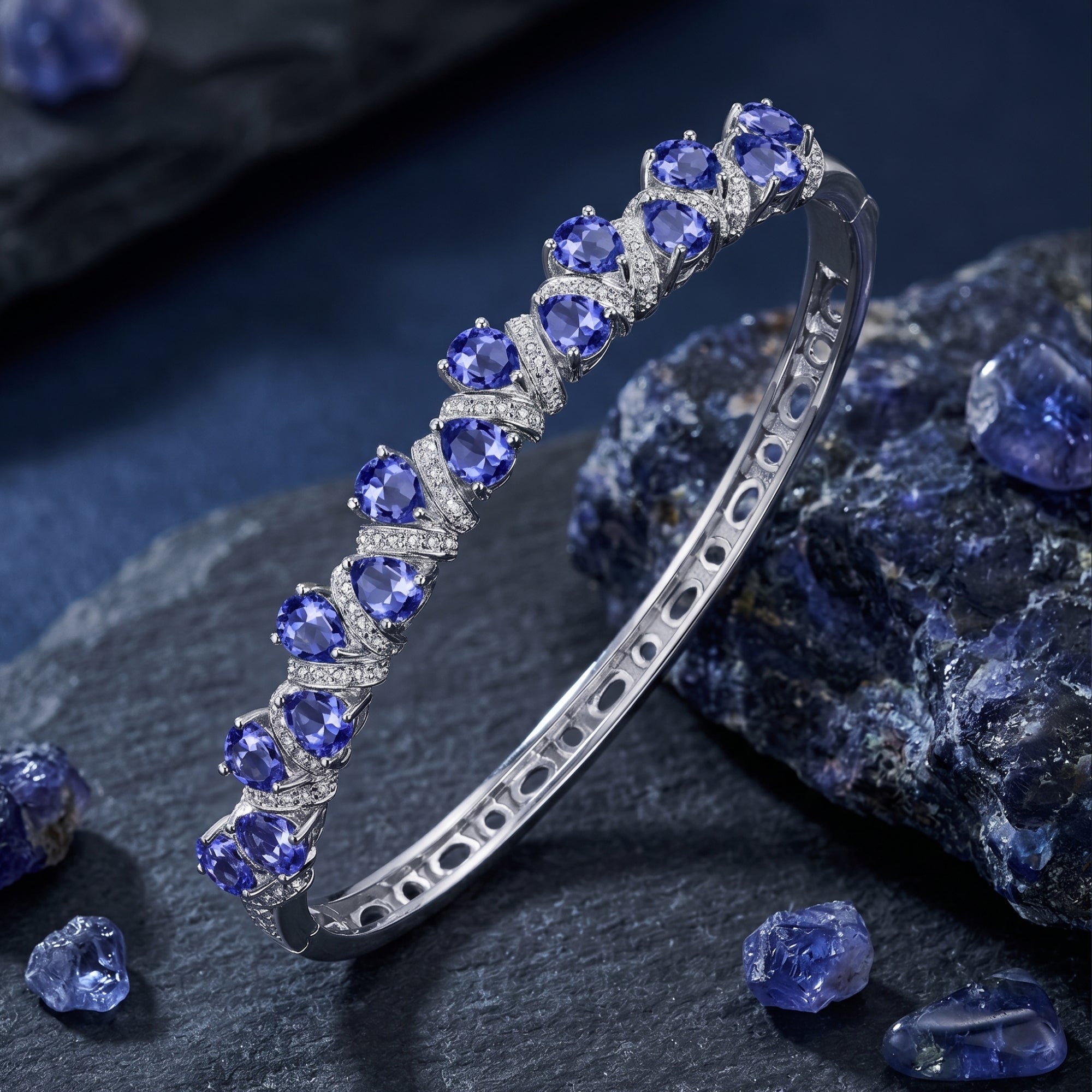 White gold pear-cut tanzanite tennis bracelet displayed on dark slate with raw gemstones.