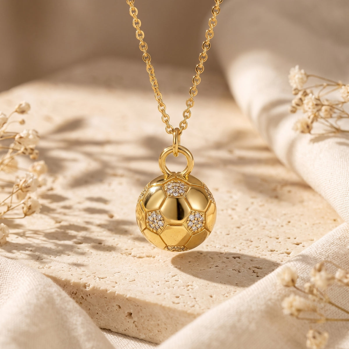 Close-up of a gold-tone soccer ball pendant with simulated-diamond accents on a chain, displayed over a stone slab.
