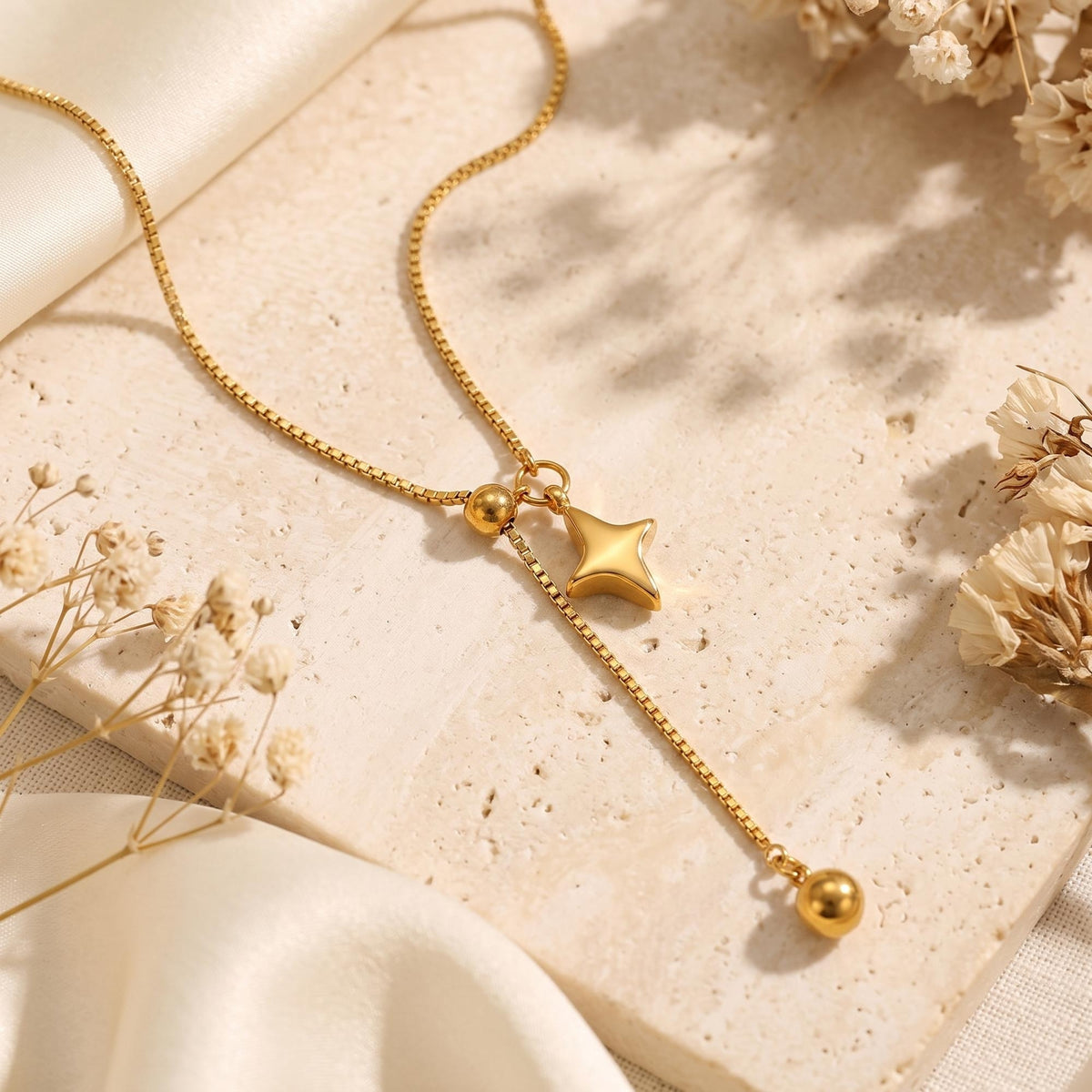Gold-tone star lariat necklace arranged flat on beige stone with cream fabric and dried flowers.