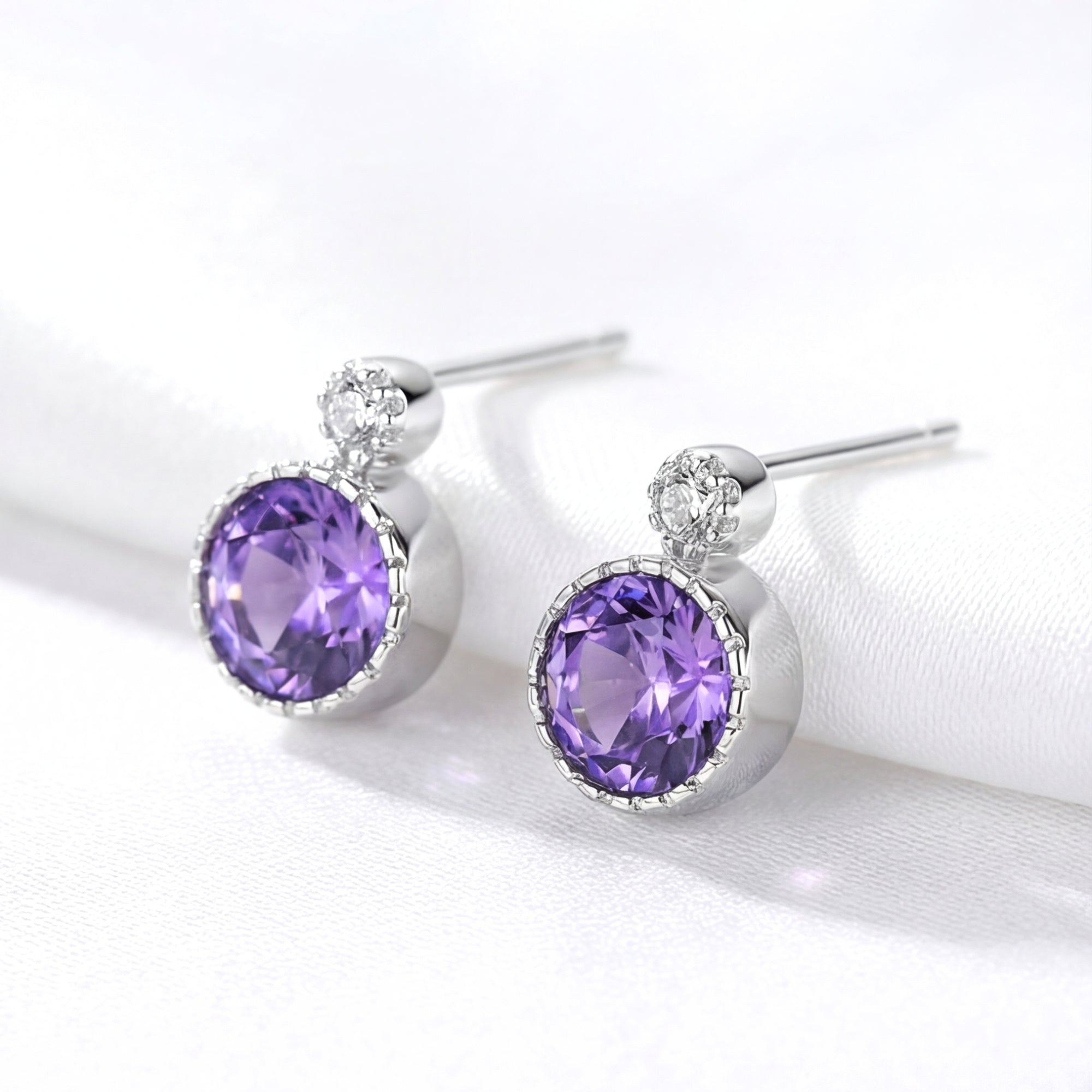 Pair of round simulated amethyst stud earrings in S925 sterling silver on a white fabric background.