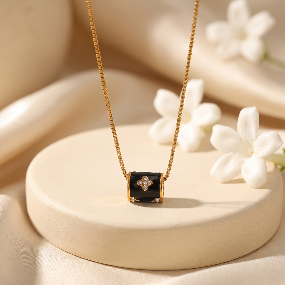 Gold-tone box chain necklace with a black enamel barrel pendant and clear stone-accented clover design on a pedestal.
