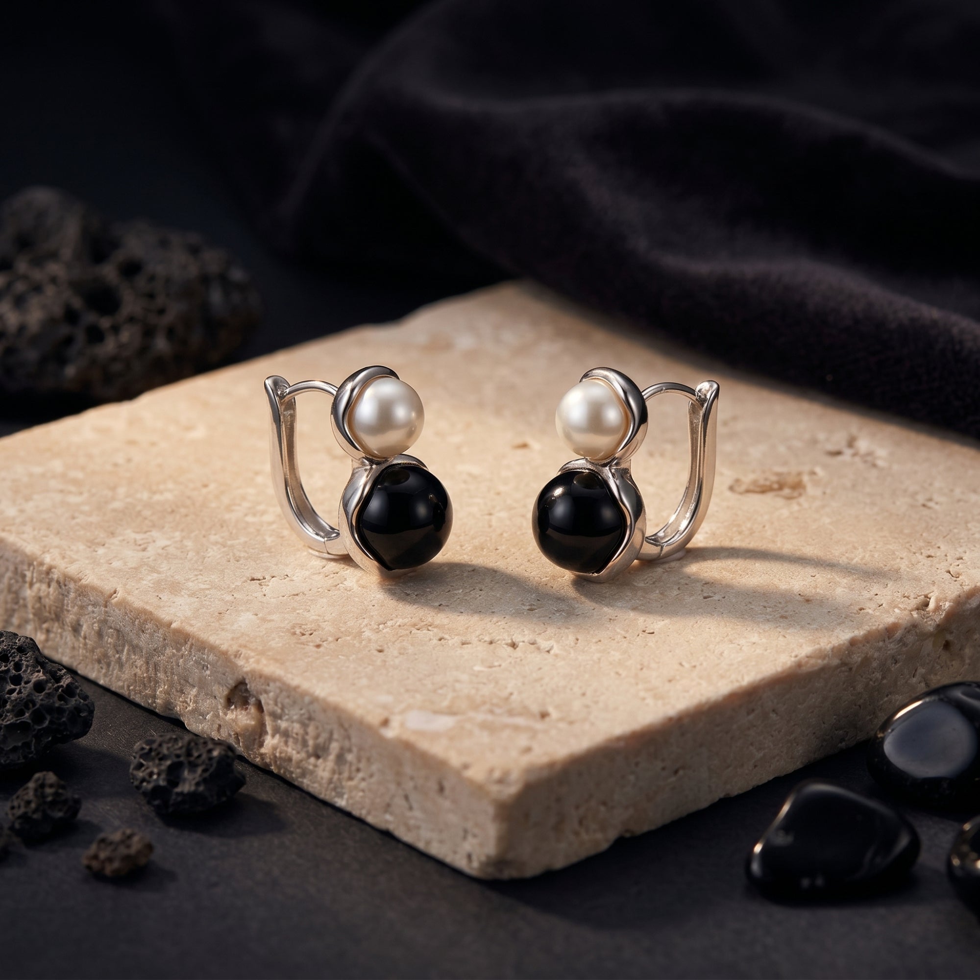 Front view of silver drop earrings with round white pearls and black onyx cabochons on a stone block with lava rocks.