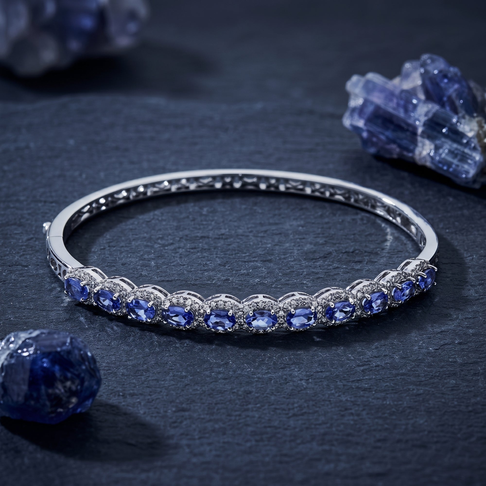 Alvira white gold bangle with 11 oval tanzanite stones displayed on a dark stone surface with raw crystals.