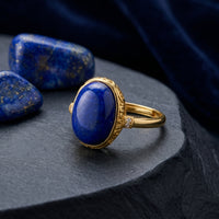 Oval lapis lazuli ring in yellow gold, styled on a dark stone surface with raw lapis lazuli gemstones.