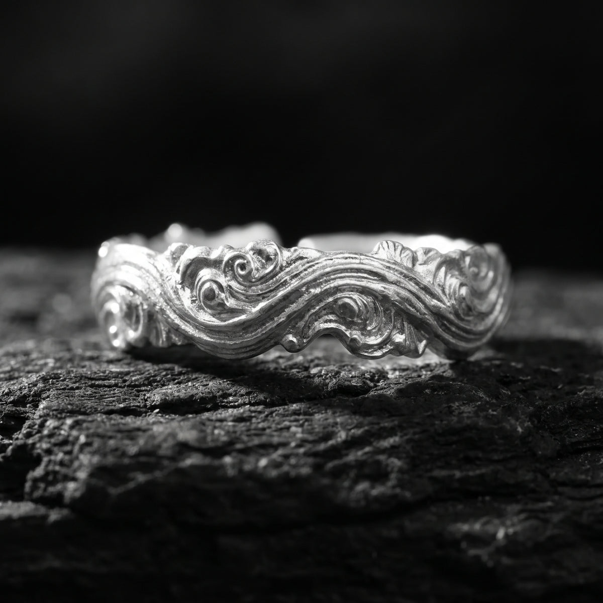 Macro front view of the fine silver wave scroll open ring resting on dark textured rock.