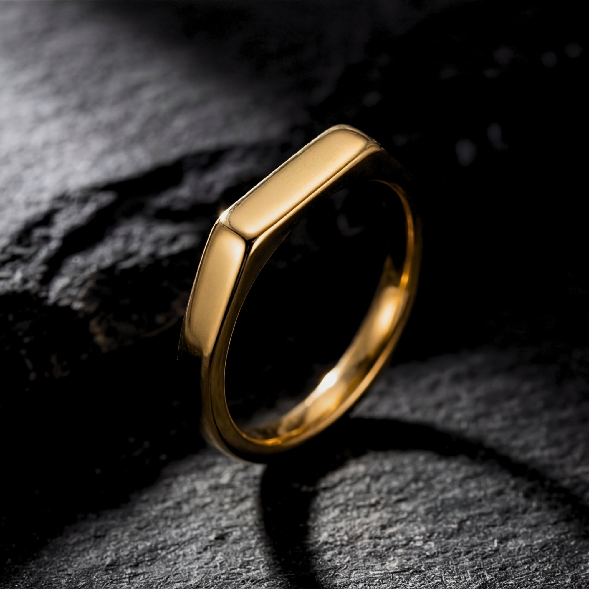 Gold stainless steel Pillar signet ring with a rectangular face on a dark textured stone background.
