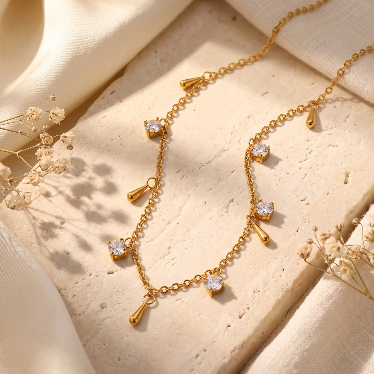 Gold-tone droplet station necklace with clear round stones and polished teardrop charms displayed on a stone slab.