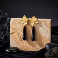 Frontal view of Calla floral drop earrings with elongated black onyx stones on a stone block, crafted in solid gold.