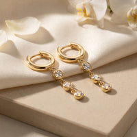 Gold moissanite drop earrings with three round stones on cream fabric and stone with white flowers.
