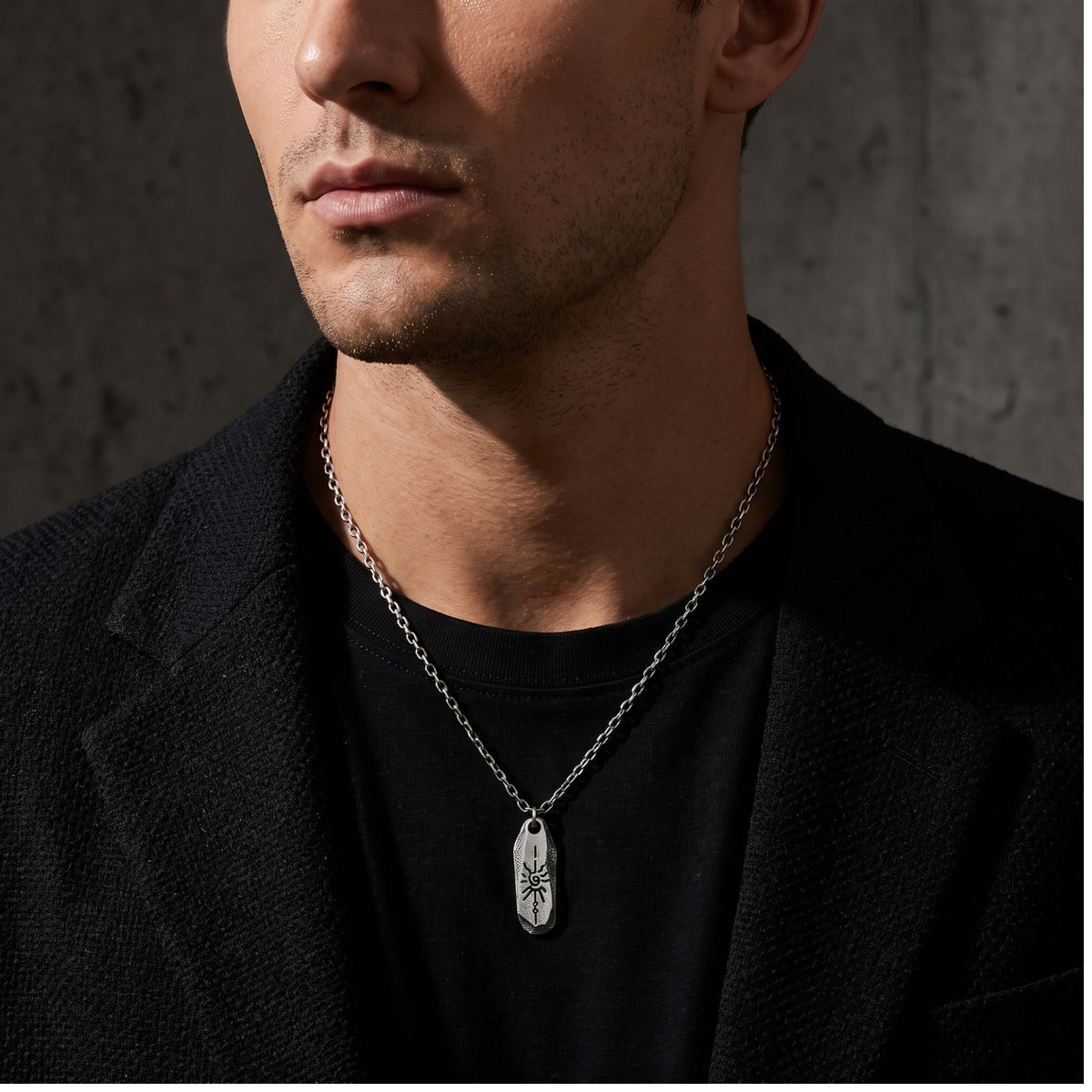 Man wearing the silver pendant necklace over a black T-shirt and beneath a dark tailored jacket.