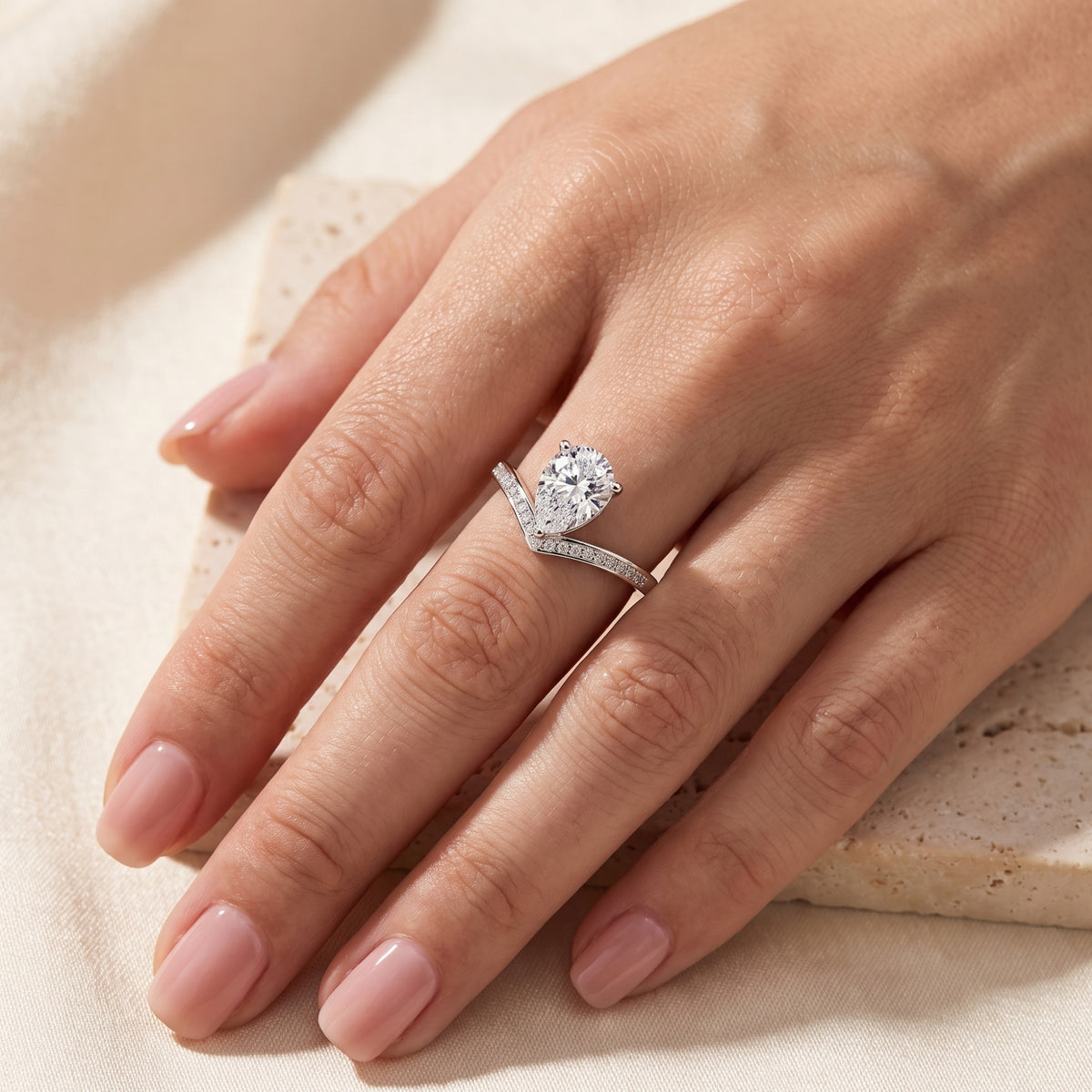 Close-up of a hand wearing a silver pear-shaped chevron ring with pave accents on a beige fabric surface.
