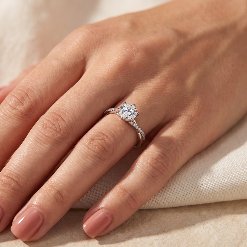 Silver crossover ring with a round clear center stone worn on a hand over cream-colored fabric.