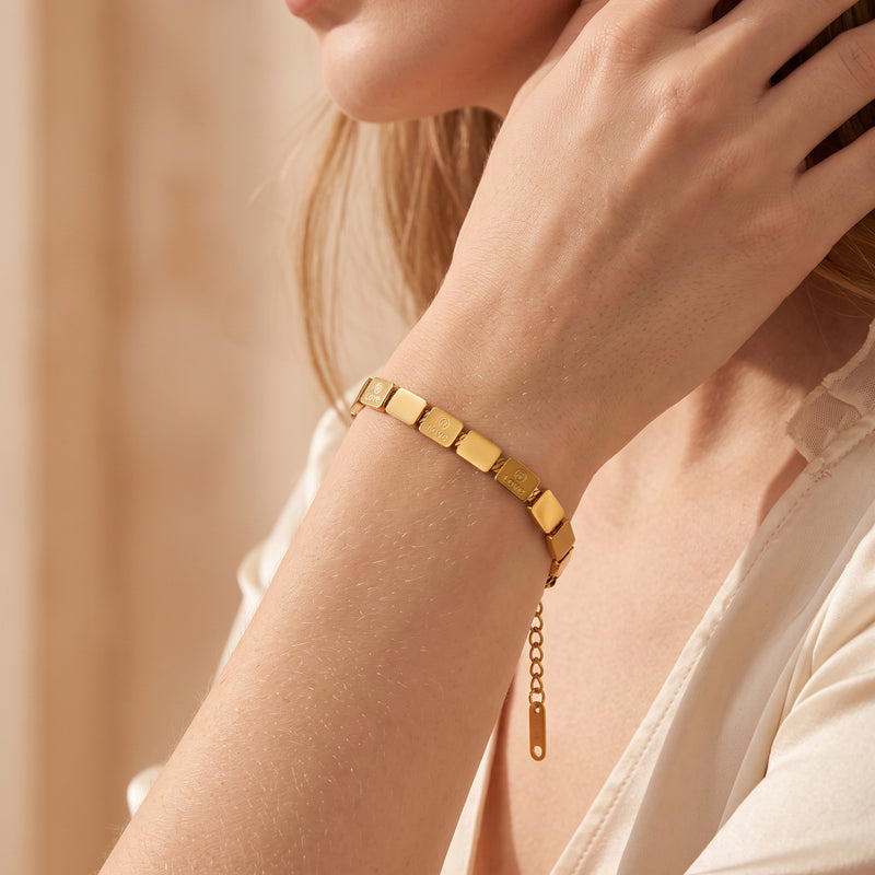 Gold-tone tile link bracelet worn on a woman's wrist, styled with a cream blouse.