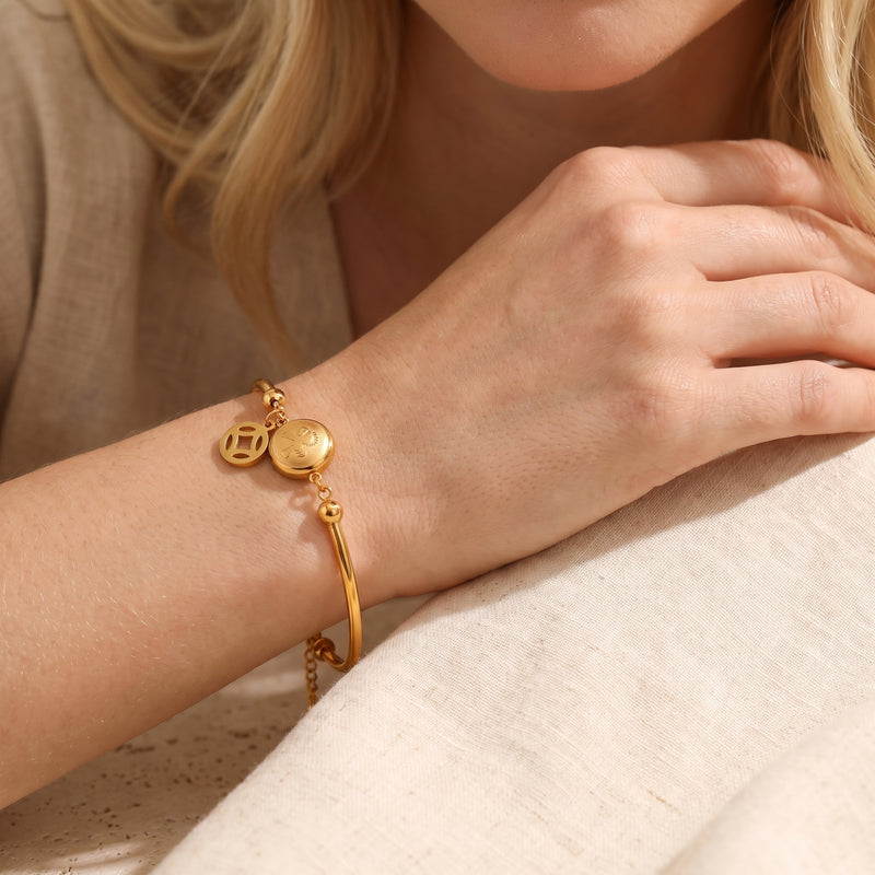 Gold-tone station bracelet worn on a wrist, showing its round medallion and openwork dangling charm over beige linen.