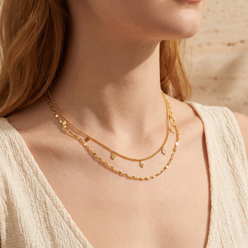 Gold-tone layered necklace with diamond-shaped charms worn on a woman's neck above a cream textured top.