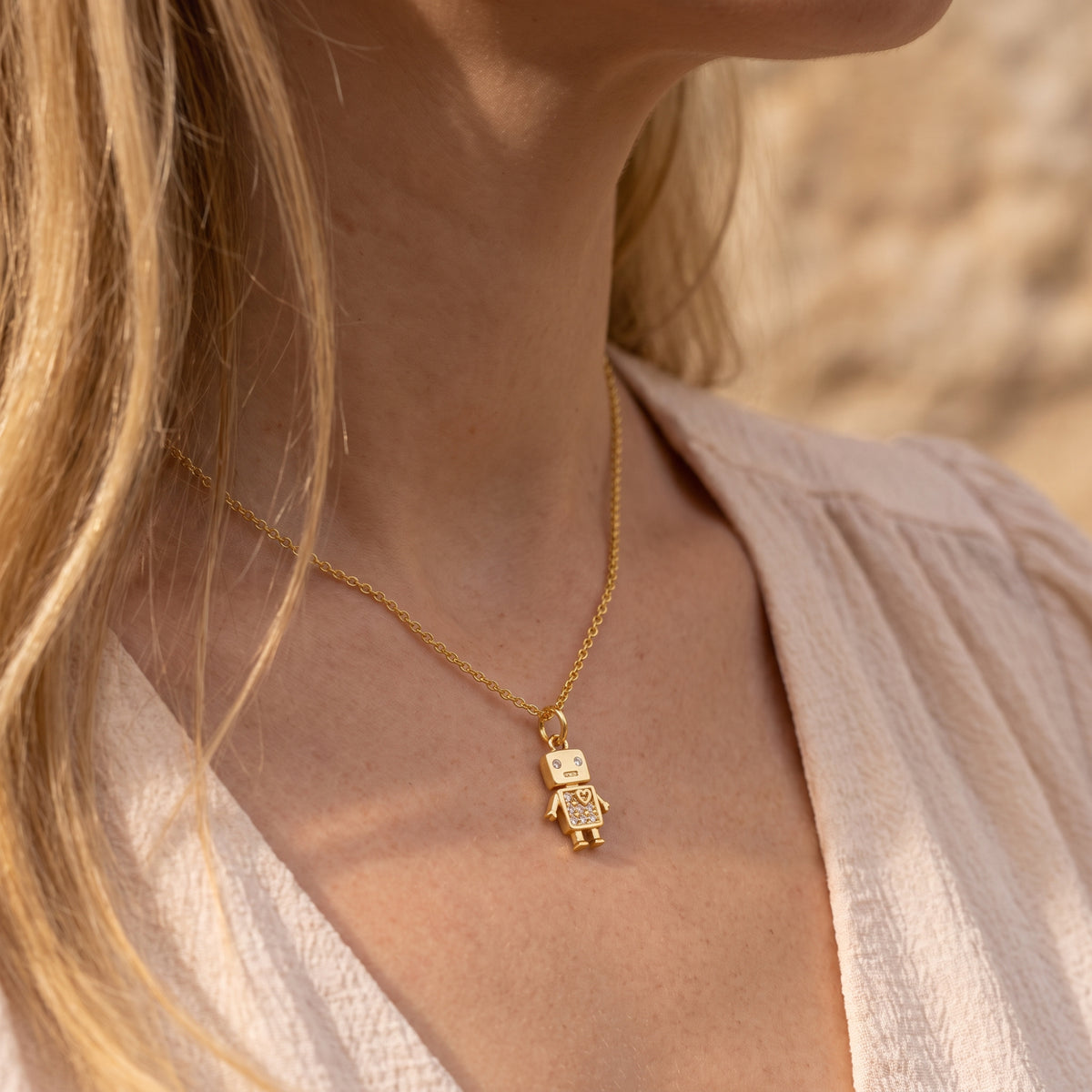 Model wearing a gold-tone robot pendant necklace with crystal details against a light cream top.