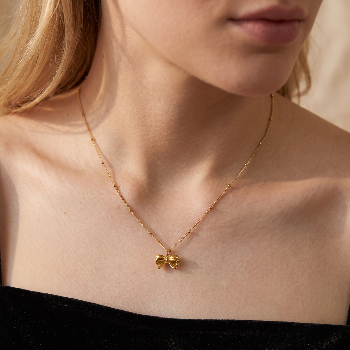 Gold-tone ribbon bow necklace on a beaded chain, worn at the neck above a black velvet top.