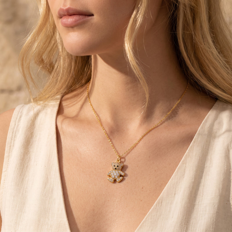Woman wearing a gold-tone crystal-paved teddy bear pendant necklace with a cream V-neck top.