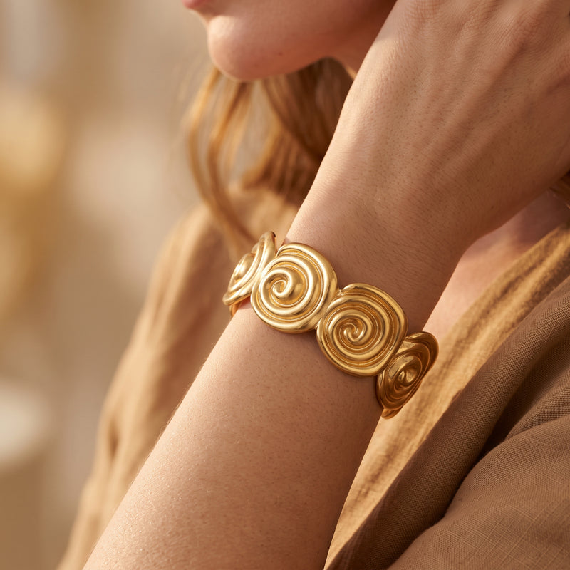 Close-up of a model wearing the gold-tone spiral motif cuff bracelet on her wrist with tan clothing behind it.