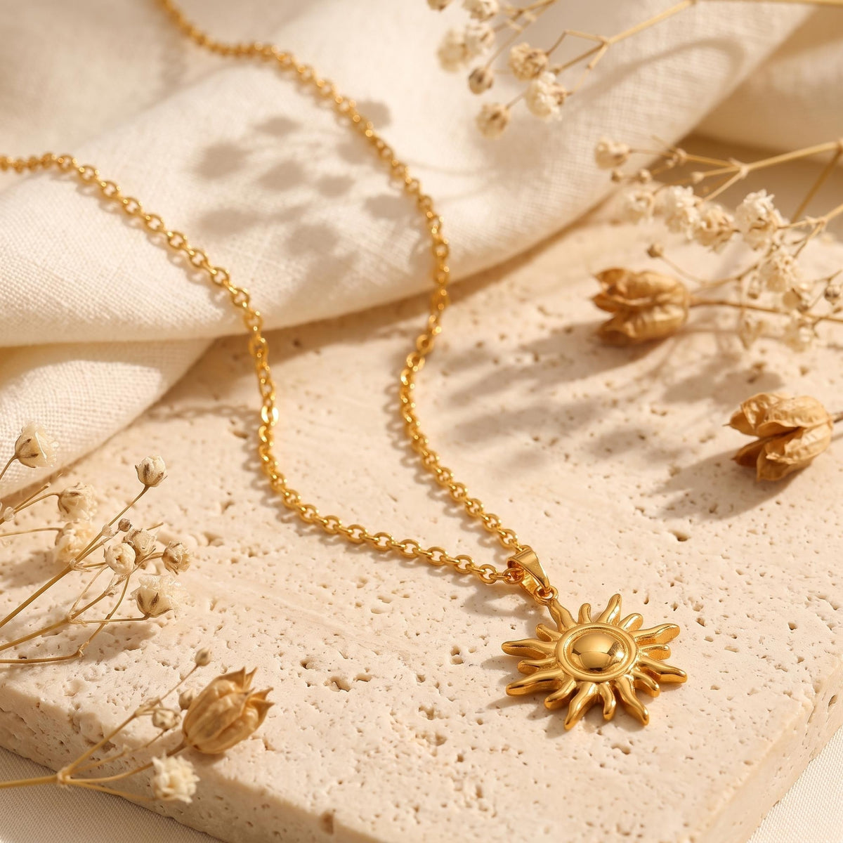 Angled view of the gold-tone sun necklace laid on a textured stone slab with cream cloth and dried flowers.