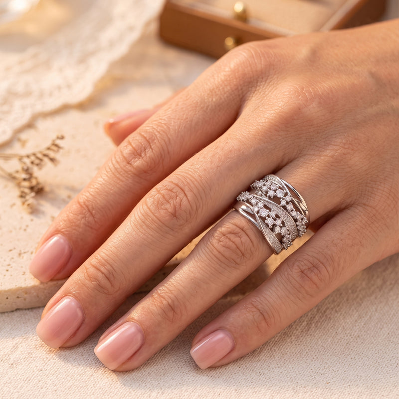 Wide silver crossover cluster ring worn on a hand, resting flat against a light textured backdrop.