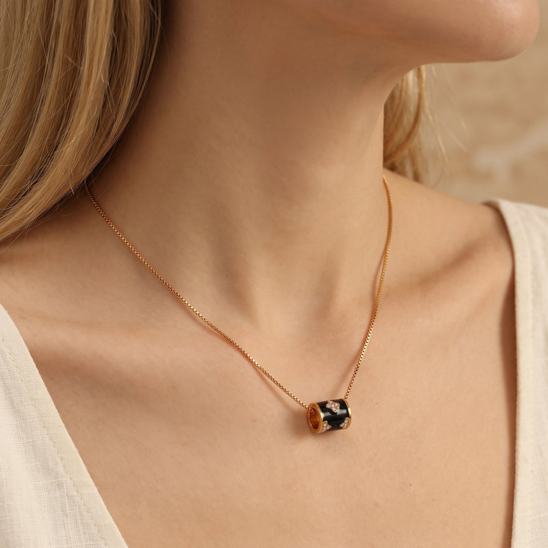 Side-view model wearing a gold-tone necklace with a black enamel barrel pendant resting near the collarbone.