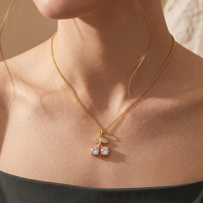 Gold-tone crystal cherry pendant necklace worn against the collarbone with a black off-shoulder top.