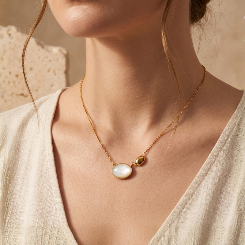 Model wearing a gold-tone necklace with an asymmetric mother-of-pearl oval and gold-tone pebble pendant.