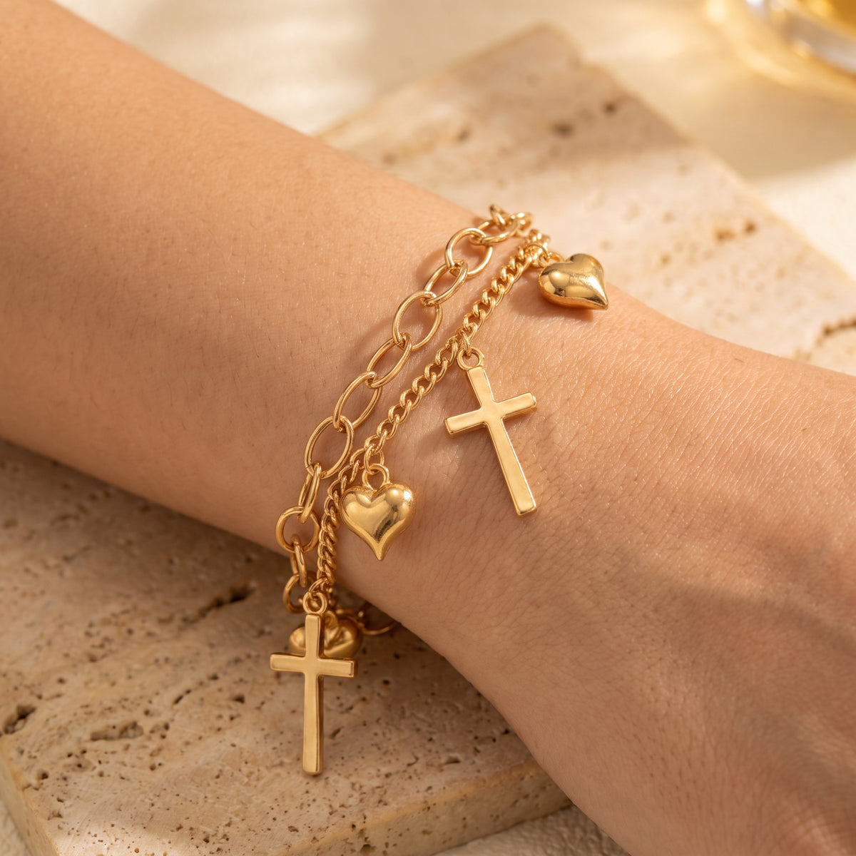 Close-up of a gold-tone double-chain bracelet with heart and cross charms worn on a wrist.