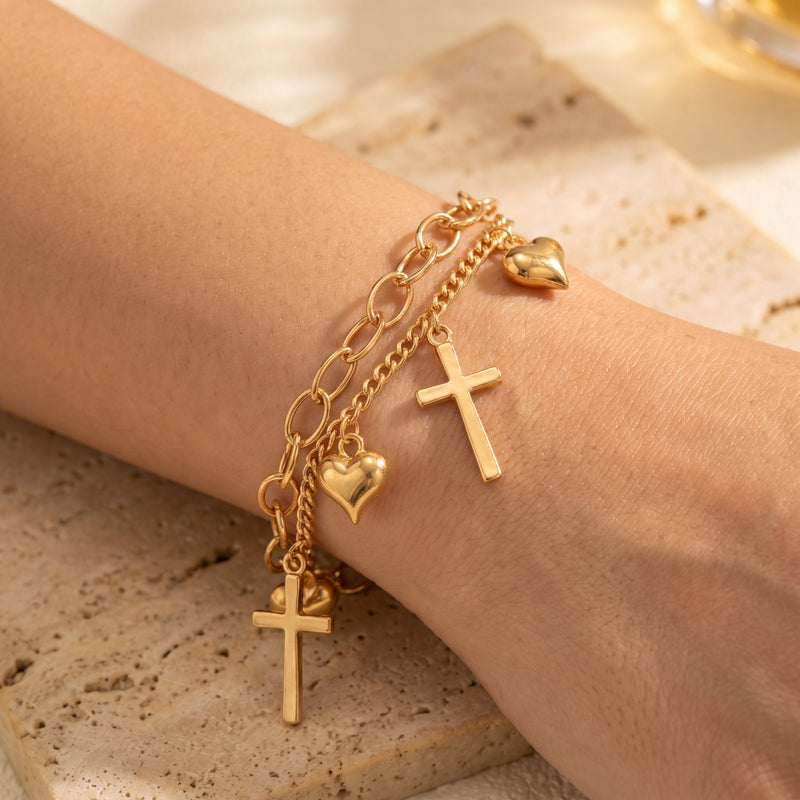 Close-up of a gold-tone double-chain bracelet with heart and cross charms worn on a wrist.